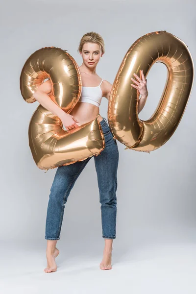 Atractiva chica en jeans y sujetador blanco posando con 20 globos dorados, en gris — Stock Photo