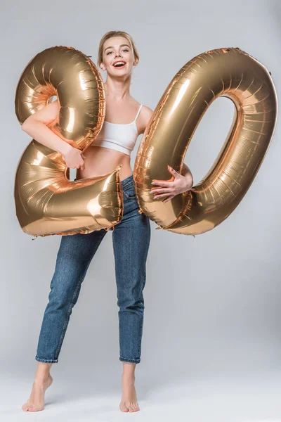 Happy girl in jeans and white bra posing with 20 golden balloons, on grey — Stock Photo