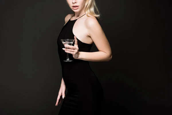 Recortado vista de la joven chica en vestido negro posando con copa de cóctel aislado en negro - foto de stock