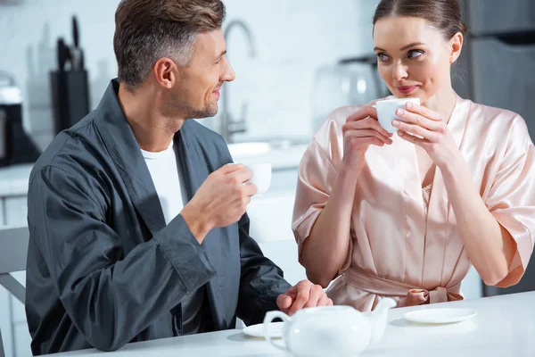 Красивая улыбающаяся взрослая пара в халатах, пьющая чай на кухне — стоковое фото