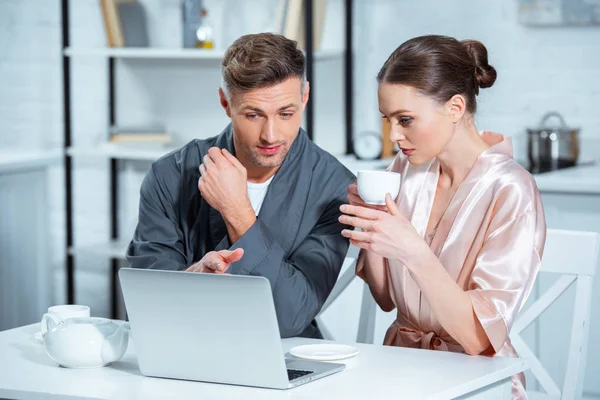 Erwachsenes Paar in Bademänteln mit Laptop beim Frühstück in der Küche — Stockfoto