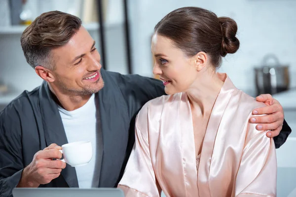 Guapo sonriente hombre con taza de té abrazando hermosa mujer en la cocina - foto de stock