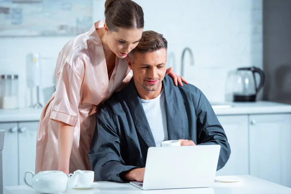 Erwachsenes Paar in Bademänteln mit Laptop beim Frühstück in der Küche — Stockfoto