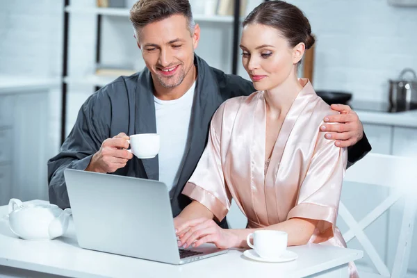 Adult couple in robes sitting at table with tea cups and using laptop during breakfast in kitchen — Stock Photo