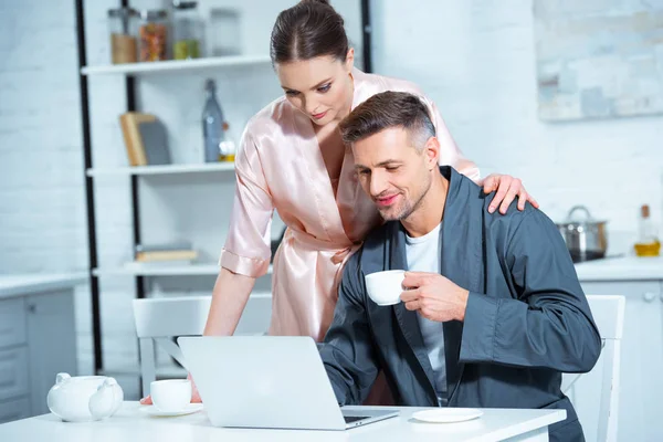 Selective focus of beautiful adult couple in robes using laptop during breakfast in kitchen — Stock Photo
