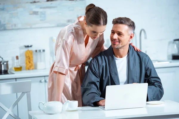 Foco seletivo do casal adulto em roupões usando laptop durante o café da manhã na cozinha — Fotografia de Stock