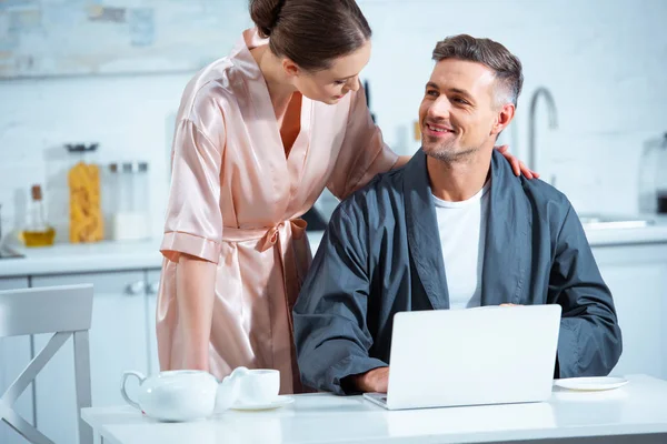 Foco seletivo do casal adulto em roupões usando laptop durante o café da manhã na cozinha — Fotografia de Stock