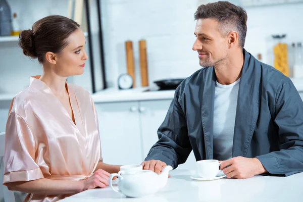 Beautiful adult couple in robes looking at each other while having tea in kitchen — Stock Photo