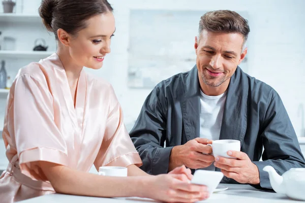 Beau couple en peignoirs en utilisant smartphone tout en prenant le thé pendant le petit déjeuner en cuisine — Photo de stock