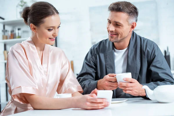 Beautiful smiling couple in robes using smartphone while having tea during breakfast in kitchen — Stock Photo