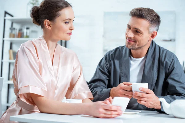 Beautiful couple in robes using smartphone while having tea during breakfast in kitchen — Stock Photo