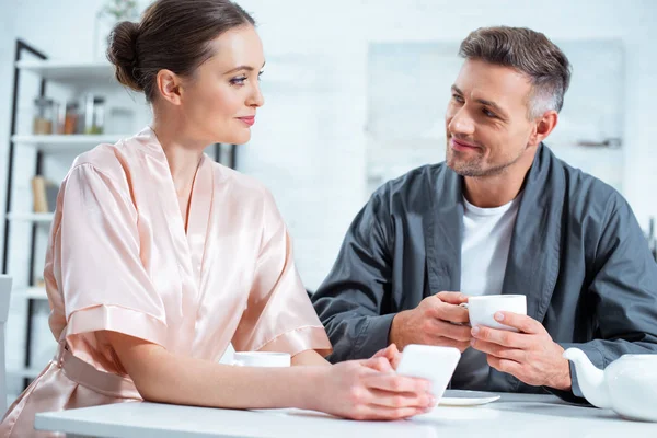 Schönes Paar in Bademänteln mit Smartphone beim Tee beim Frühstück in der Küche — Stockfoto