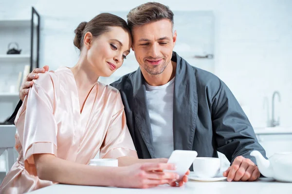 Beau couple souriant en peignoirs en utilisant smartphone tout en prenant le thé pendant le petit déjeuner dans la cuisine — Photo de stock