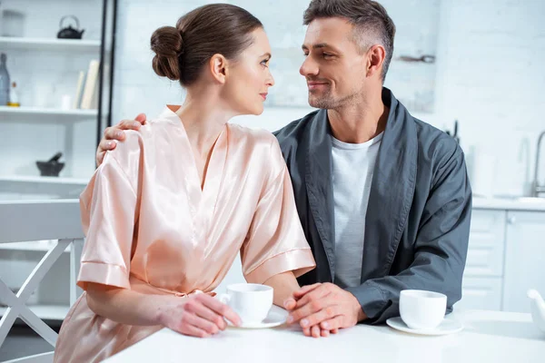 Beautiful adult couple in robes looking at each other while having tea in kitchen — Stock Photo