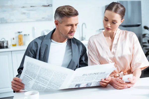 Foco seletivo de casal em robes lendo jornal de negócios na cozinha — Fotografia de Stock