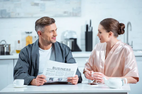 Чоловік в халаті читає бізнес-газету, а жінка п'є чай під час сніданку на кухні — стокове фото