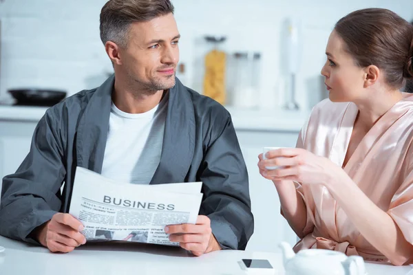 Чоловік в халаті читає бізнес-газету, а жінка п'є чай під час сніданку на кухні — стокове фото