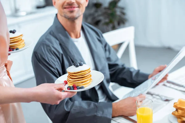 Vista ritagliata di donna che tiene piatti con frittelle mentre l'uomo legge il giornale durante la colazione in cucina — Foto stock