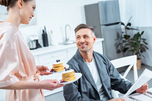 Woman holding plates with pancakes while smiling man reading newspaper during breakfast in kitchen — Stock Photo