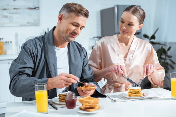 Enfoque selectivo del marido y la esposa en túnicas durante el desayuno con panqueques y jugo de naranja en la cocina - foto de stock