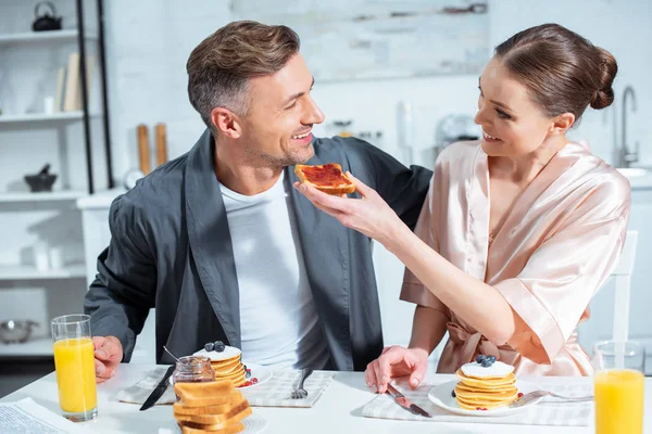 Sonriente pareja de adultos en túnicas durante el desayuno con panqueques y jugo de naranja en la cocina - foto de stock