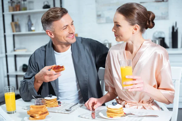 Marido y mujer en batas durante el desayuno con panqueques y jugo de naranja en la cocina - foto de stock