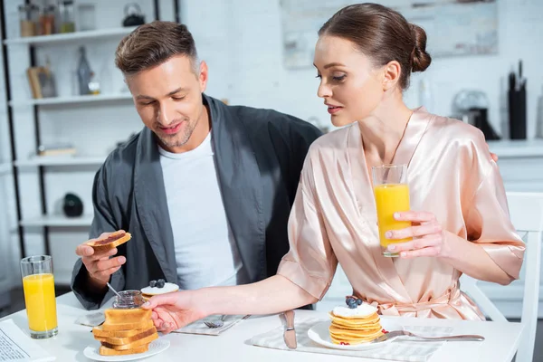 Selective focus of husband and wife in robes during breakfast with pancakes and orange juice in kitchen — Stock Photo