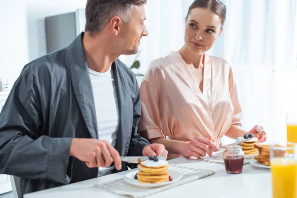 Hermosa pareja en túnicas durante el desayuno con panqueques y jugo de naranja en la cocina - foto de stock