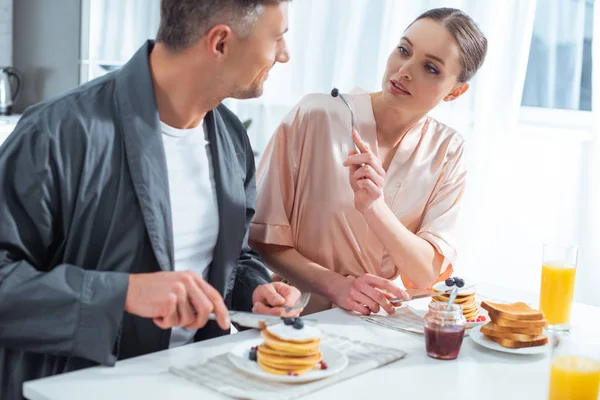 Beau couple en peignoirs pendant le petit déjeuner avec crêpes et jus d'orange en cuisine — Photo de stock