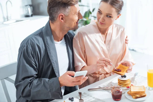 Beautiful adult couple in robes using smartphone while having breakfast in kitchen — Stock Photo