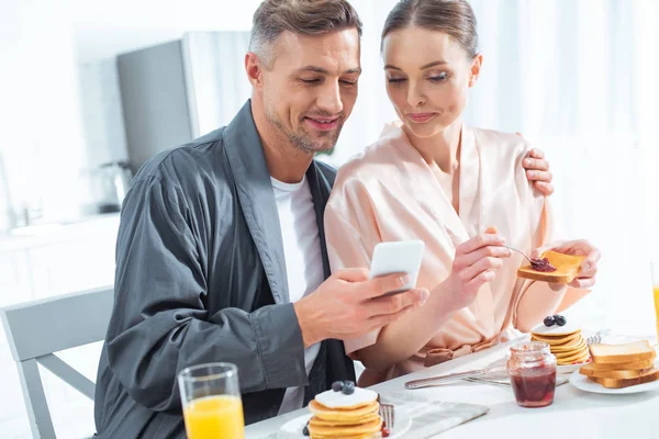 Красивая взрослая пара в халатах с помощью смартфона во время завтрака на кухне — стоковое фото