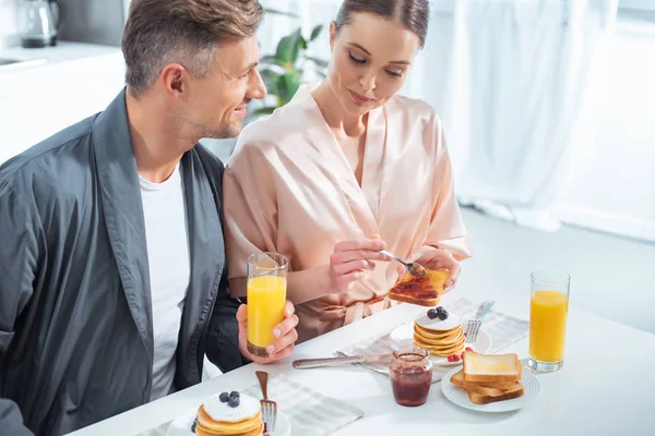 Foco seletivo de belo casal em roupões durante o café da manhã com panquecas e suco de laranja na cozinha — Fotografia de Stock