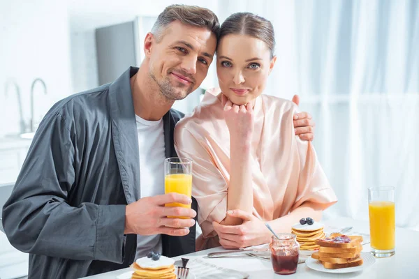 Красивый мужчина держит апельсиновый сок и обнимает женщину во время завтрака утром — стоковое фото