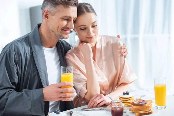 Красивый мужчина держит апельсиновый сок и обнимает улыбающуюся женщину во время завтрака утром — стоковое фото