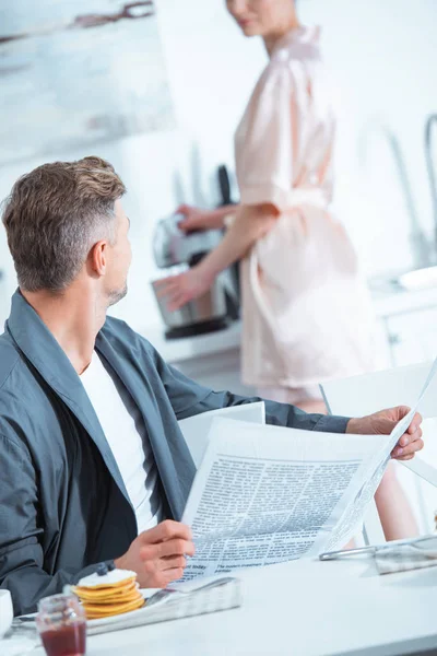 Foyer sélectif de l'homme lisant le journal tandis que la femme cuisine sur le fond le matin — Photo de stock