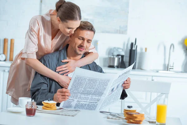 Frau in Robe umarmt gutaussehenden Mann beim Frühstück in der Küche mit Zeitung — Stockfoto
