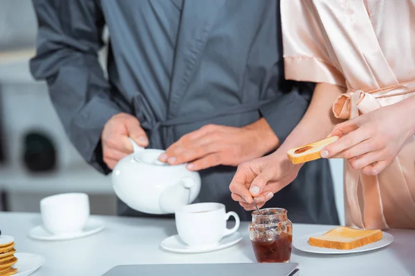 Vista recortada del hombre sirviendo té mientras la mujer prepara tostadas con mermelada durante el desayuno - foto de stock