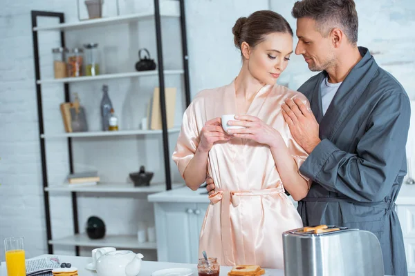 Hombre guapo en bata abrazando hermosa mujer con taza de té en la cocina durante el desayuno - foto de stock