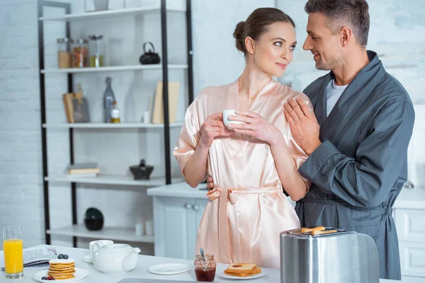 Handsome man in robe embracing beautiful woman with cup of tea in kitchen during breakfast — Stock Photo