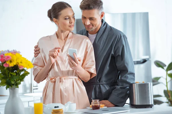 Beau couple adulte en peignoirs en utilisant smartphone pendant le petit déjeuner dans la cuisine — Photo de stock