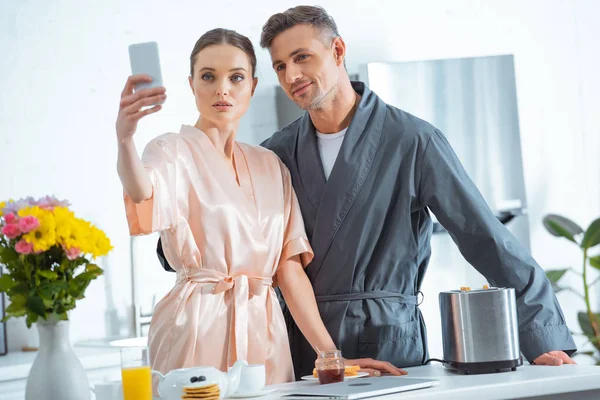 Красивая взрослая пара в халатах делает селфи на смартфоне во время завтрака на кухне — стоковое фото