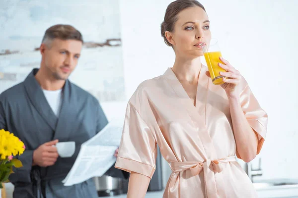 Красива жінка в халаті п'є апельсиновий сік, коли чоловік читає газету на кухні — стокове фото