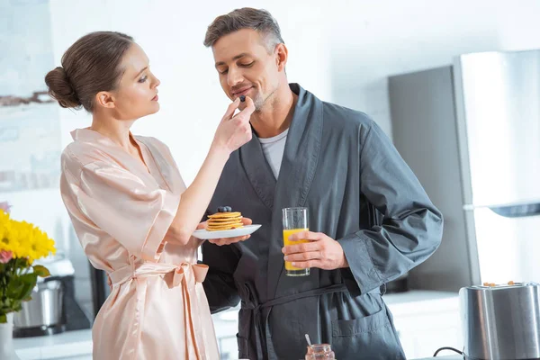Beau couple en peignoirs avec jus d'orange et crêpes pendant le petit déjeuner en cuisine — Stock Photo