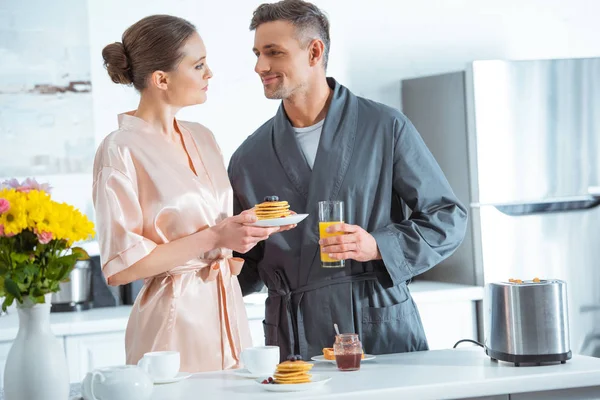 Schönes Paar in Bademänteln mit Orangensaft und Pfannkuchen beim Frühstück in der Küche — Stockfoto