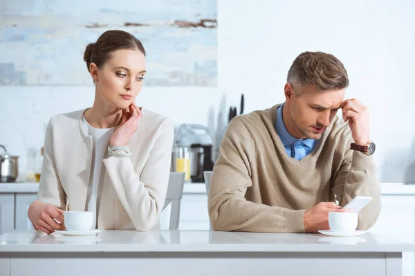 Dissatisfied man using smartphone and ignoring woman during breakfast in kitchen — Stock Photo