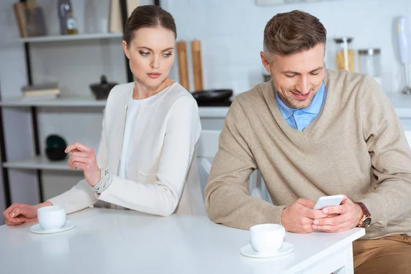 Smiling man using smartphone and ignoring woman during breakfast in kitchen — Stock Photo