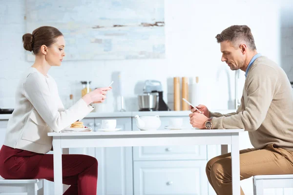 Adult couple using smartphones and ignoring each other during breakfast in kitchen — Stock Photo