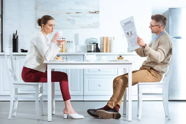 Mujer insatisfecha bebiendo té mientras el hombre lee el periódico de negocios durante el desayuno en la cocina - foto de stock