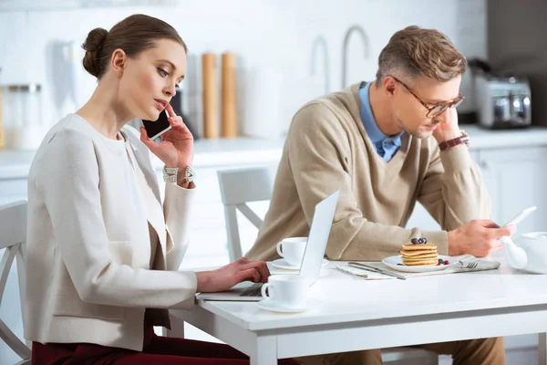 Pareja adulta sentada en la mesa, usando teléfonos inteligentes e ignorándose durante el desayuno - foto de stock