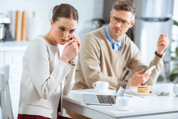 Dissatisfied man looking at woman secretly talking on smartphone during breakfast — Stock Photo